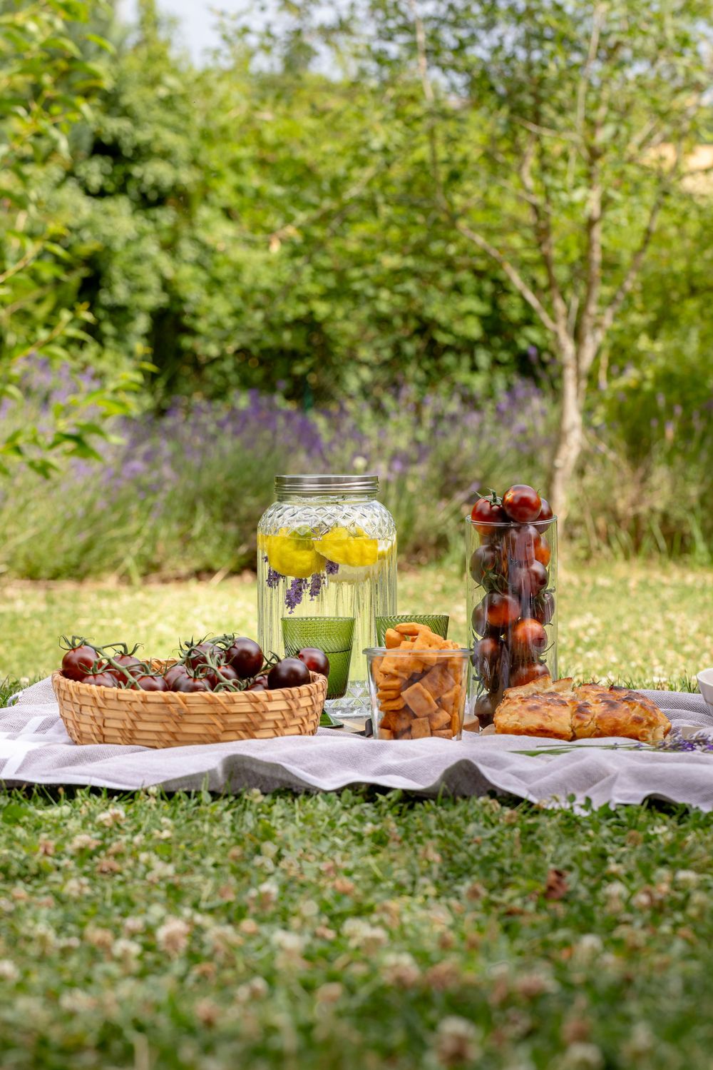 A picnic spread on grass with lemonade, cherries in a basket, cookies, and pastries, set in a lush green garden.