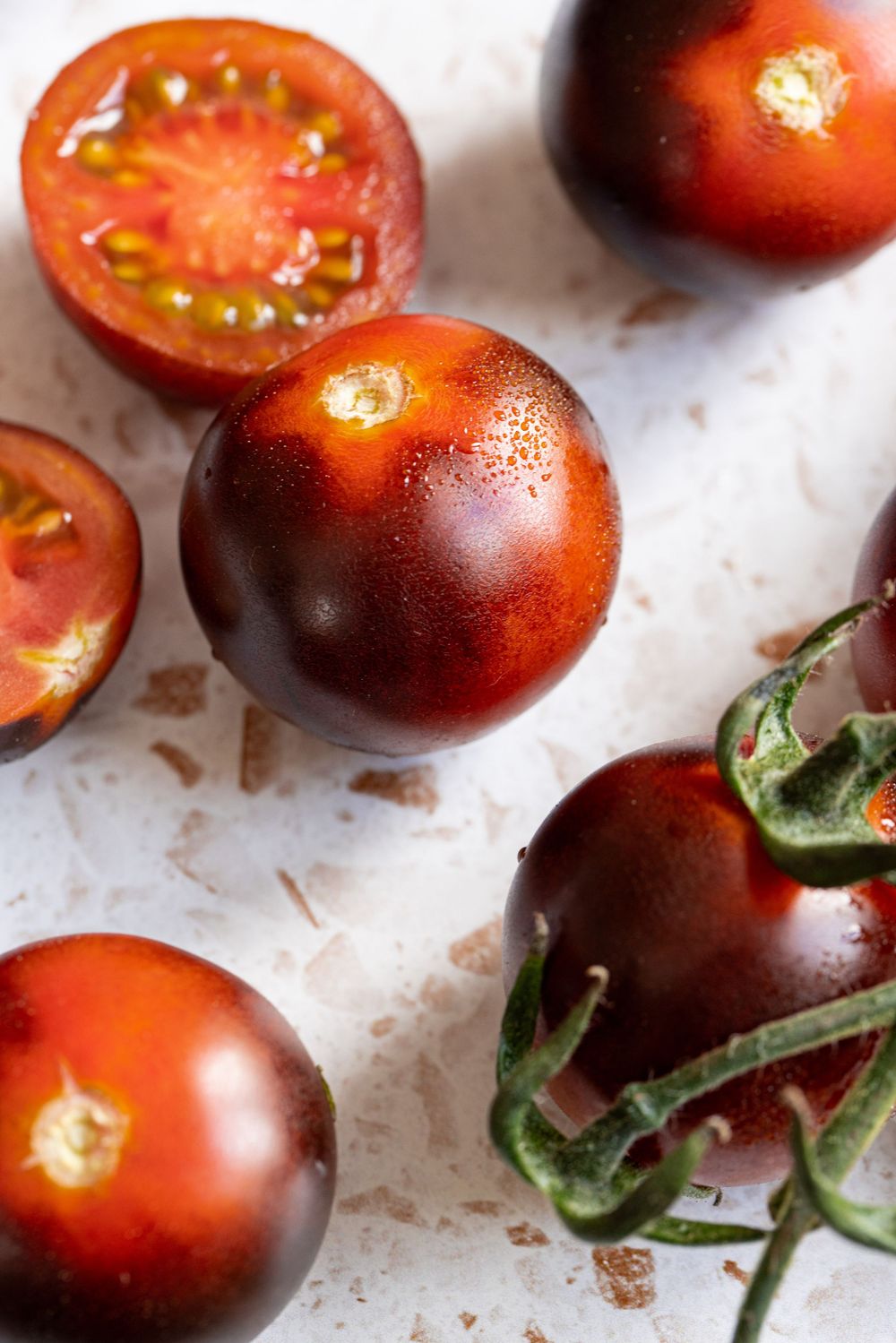 Dark purple-red heirloom cherry tomatoes, some halved, on a white surface showing their vibrant red interior.