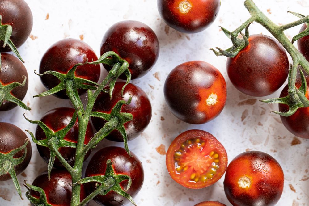 Dark purple cherry tomatoes on vine, one halved showing red interior, arranged on white surface.