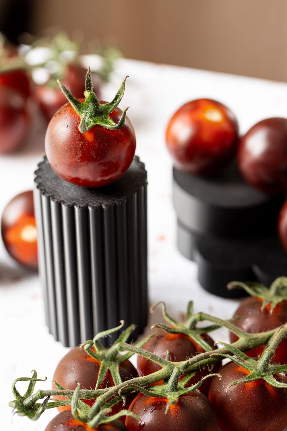 Dark red cherry tomatoes on vine displayed with black cylindrical containers on white surface.