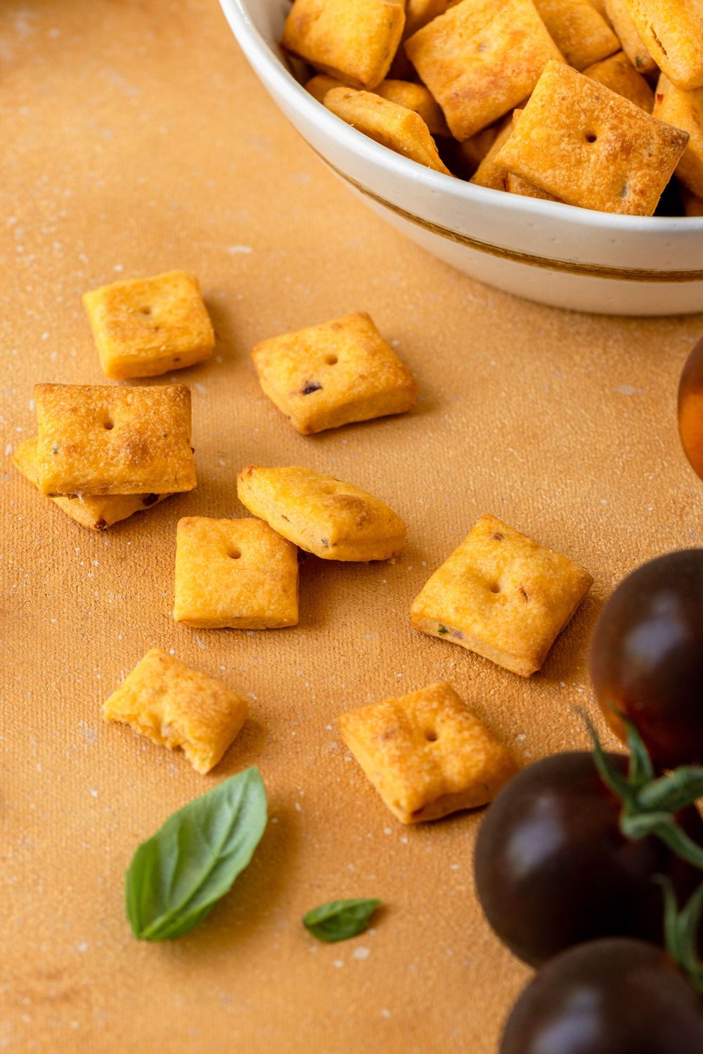 Golden square crackers scattered near a white bowl, fresh basil leaves, and dark tomatoes on an orange surface.