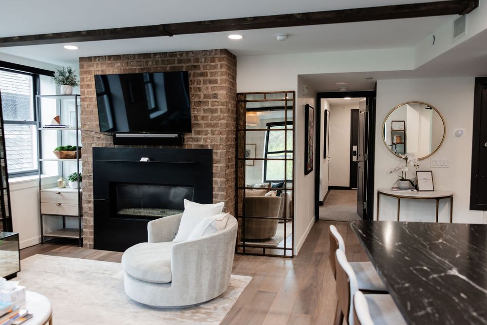 Modern living room with brick fireplace, wall-mounted TV, round accent chair, and open hallway with gold mirror.