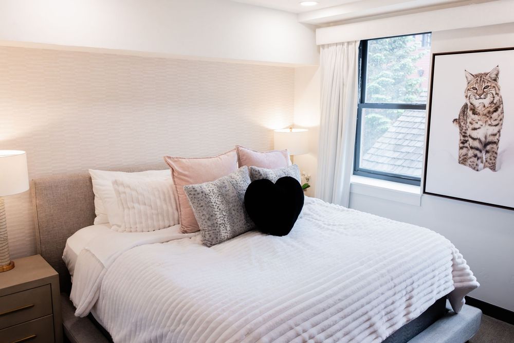 Bright bedroom with white bedding, pink and gray pillows, black heart pillow, bedside lamp, and framed cat art by window.