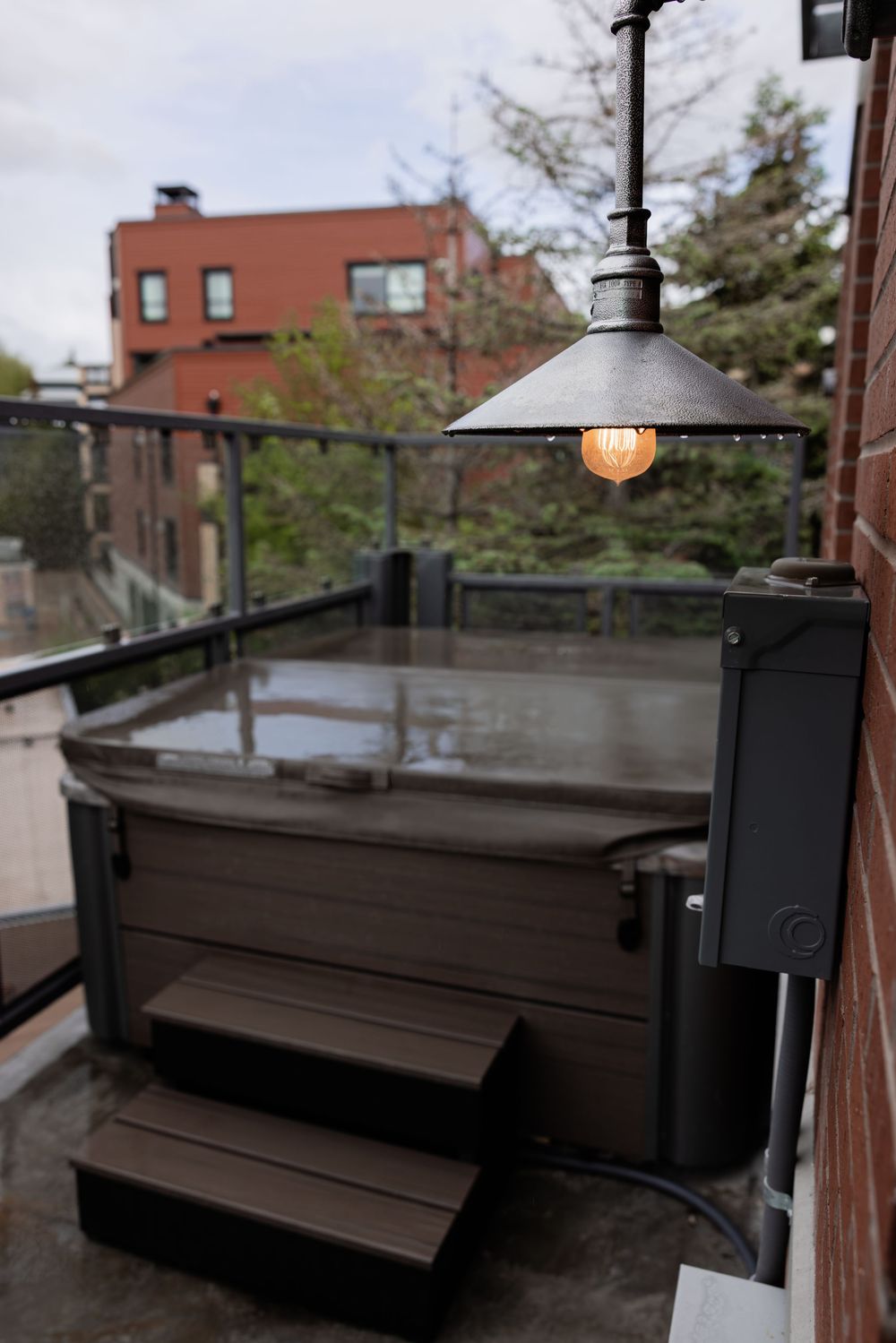 Outdoor rooftop hot tub on a balcony with a vintage pendant light, wood steps, and urban building views.