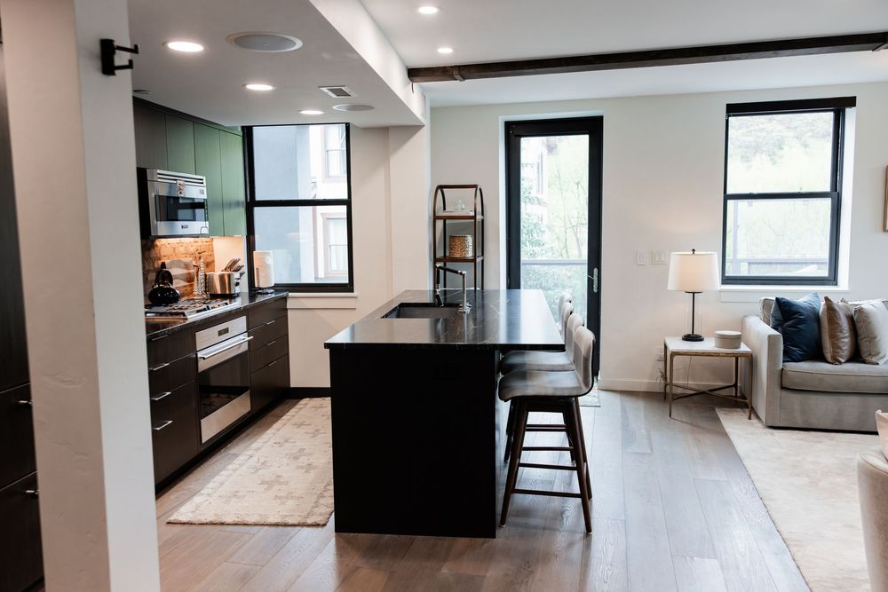 Modern open-concept kitchen with dark island, bar stools, stainless appliances, hardwood floors, and bright living area.