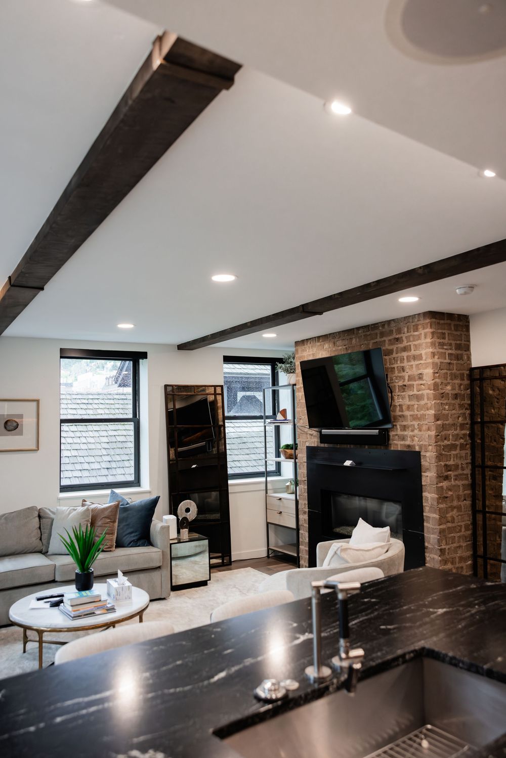 Modern living room with exposed wood beams, brick fireplace, mounted TV, and black marble kitchen island.