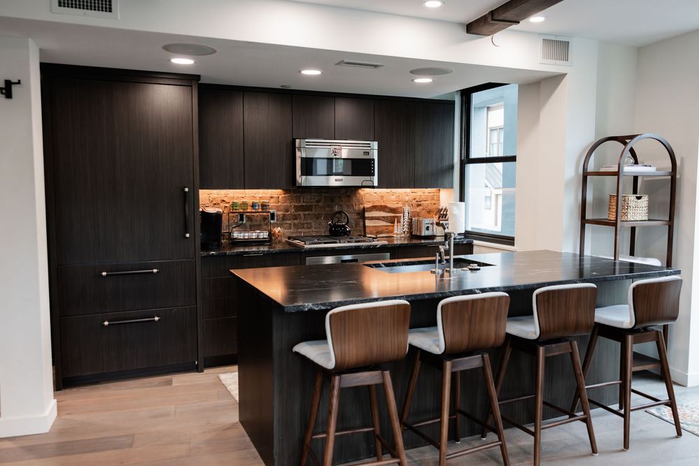 Modern kitchen with dark cabinets, black granite island, bar stools, stainless steel appliances, and warm under-cabinet lighting.