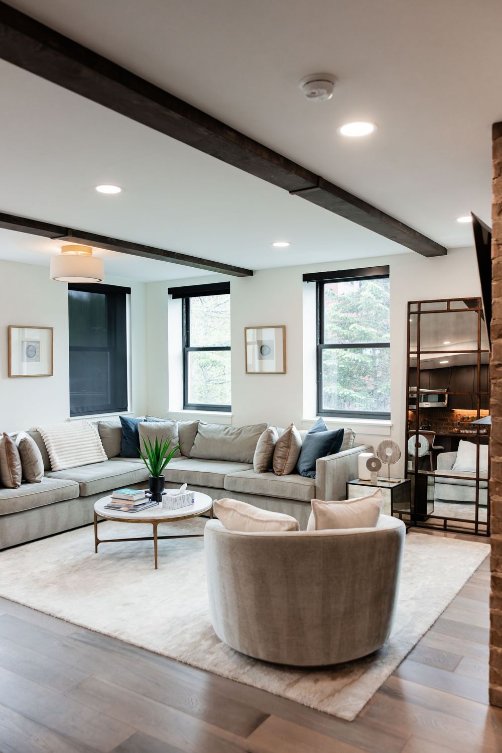 Modern living room with gray sectional sofa, round swivel chair, wood beam ceiling, and hardwood floors on a cream area rug.