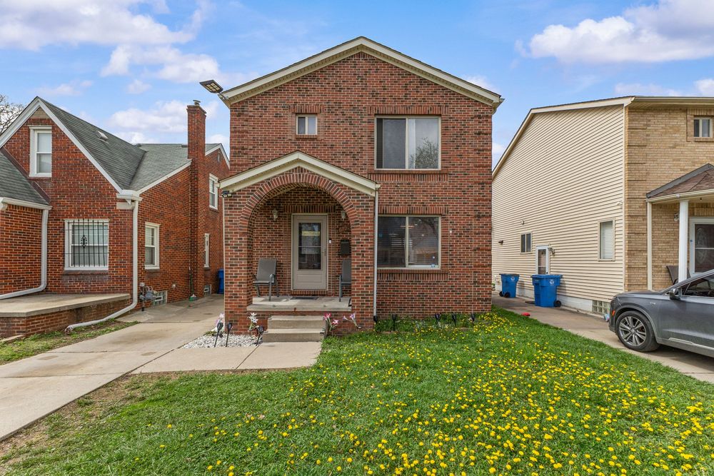 Classic brick two-story home with covered front porch, green lawn, and yellow wildflowers in urban neighborhood.
