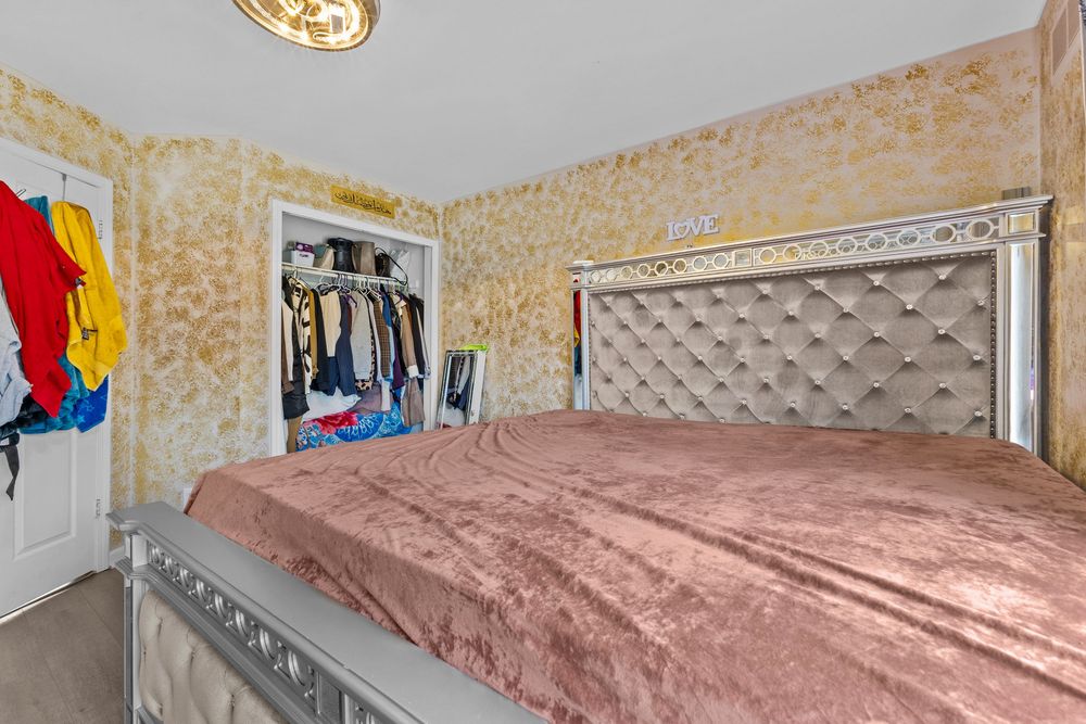 Cozy bedroom with tufted silver headboard, pink bedding, closet, and decorative gold patterned wallpaper.