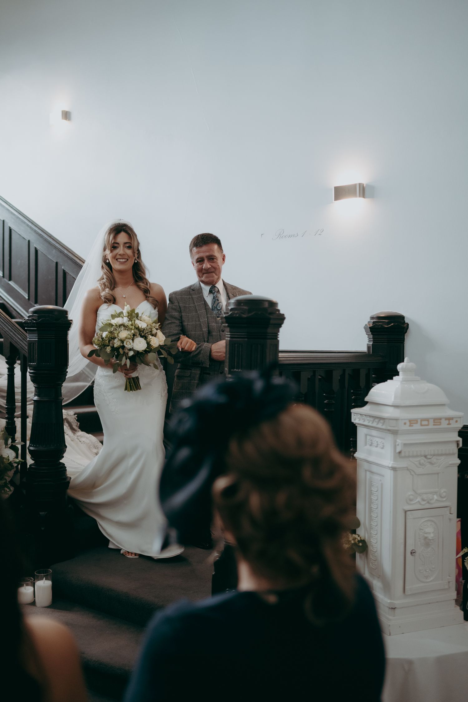 Bride in white gown descends staircase with bouquet, escorted by man in suit, at elegant wedding venue.