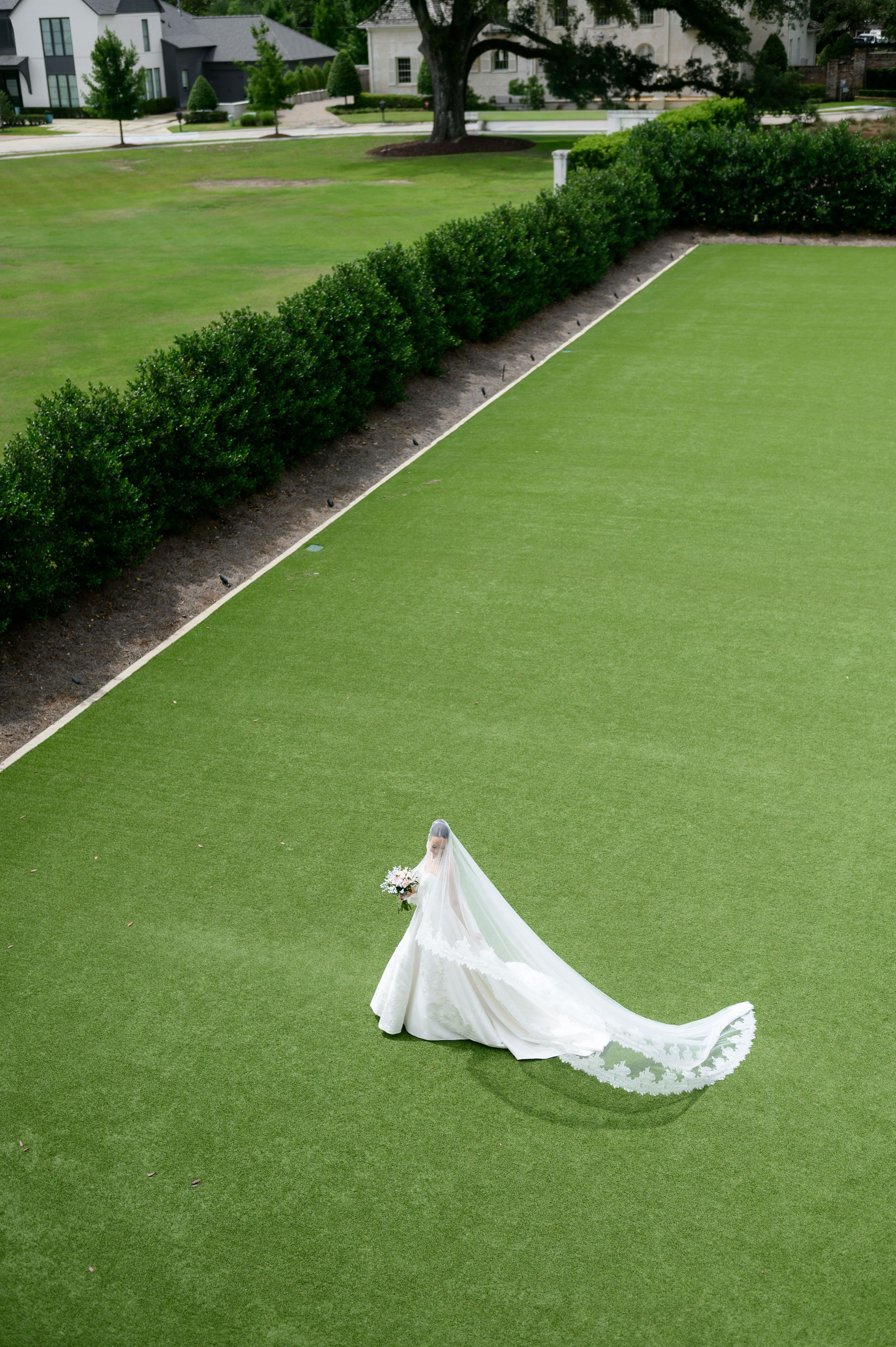 Aerial view of a bride in a white gown with long veil and train walking across a lush green lawn.