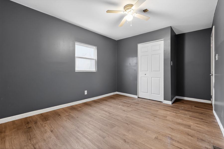 Bright bedroom with gray walls, hardwood floors, white closet door, and ceiling fan with light fixture.
