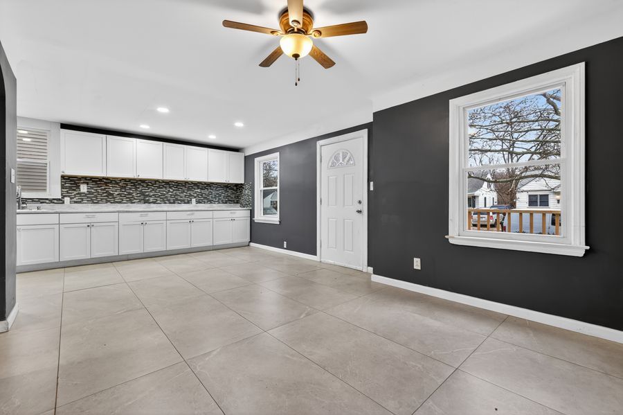 Modern kitchen with white cabinets, mosaic backsplash, large tile floors, ceiling fan, and bright window with outdoor view.