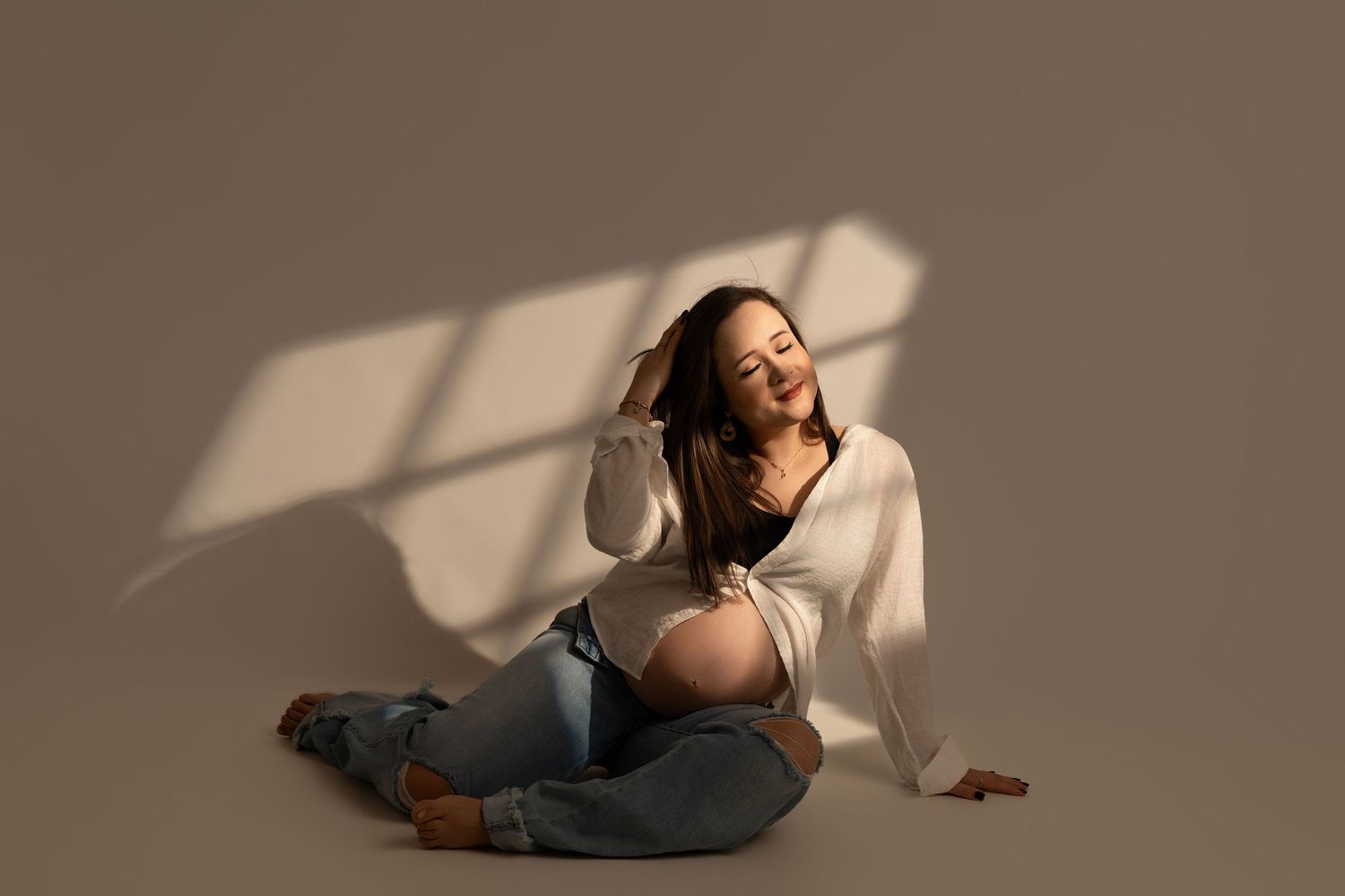 Pregnant woman in elegant white gown holding baby bump, posing gracefully beside flowing white curtains in a bright studio.