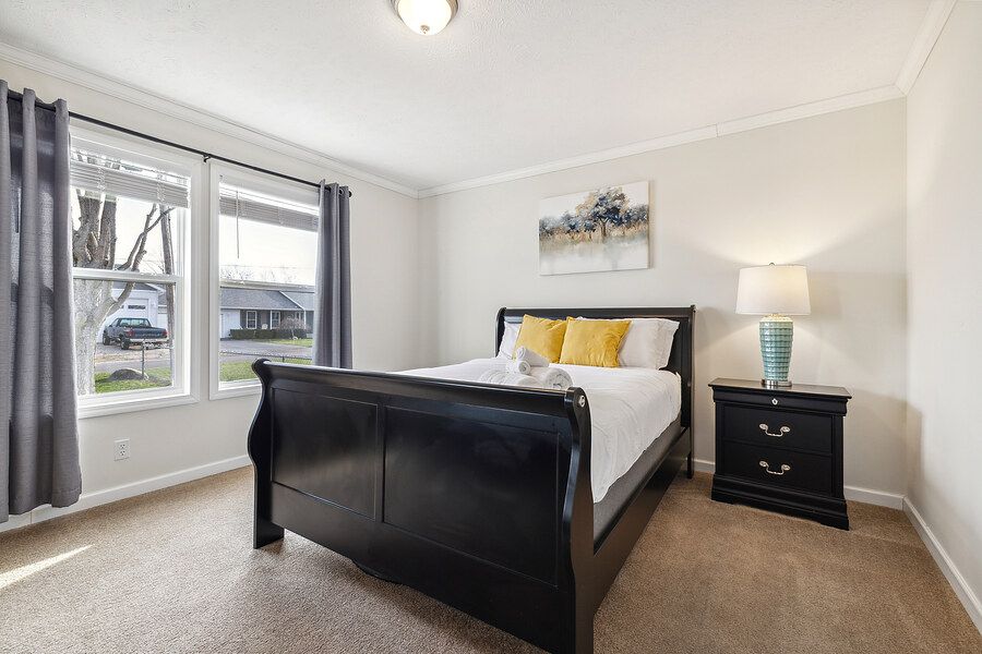 Bright bedroom with black sleigh bed, yellow accent pillows, nightstand with lamp, and large window with gray curtains.