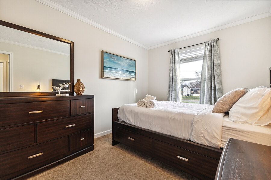 Cozy bedroom with dark wood dresser, mirror, queen bed with storage, neutral carpet, and a bright window with curtains.