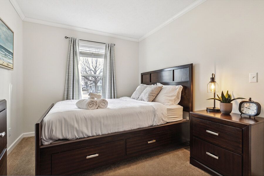 Elegant bedroom with dark wood queen bed frame, white bedding, nightstand with lamp, and neutral carpet flooring.