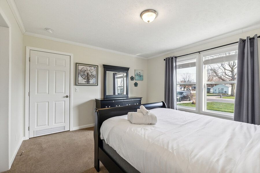 Bright bedroom with a large bed, dark wood furniture, carpeted floors, crown molding, and a window with gray curtains.