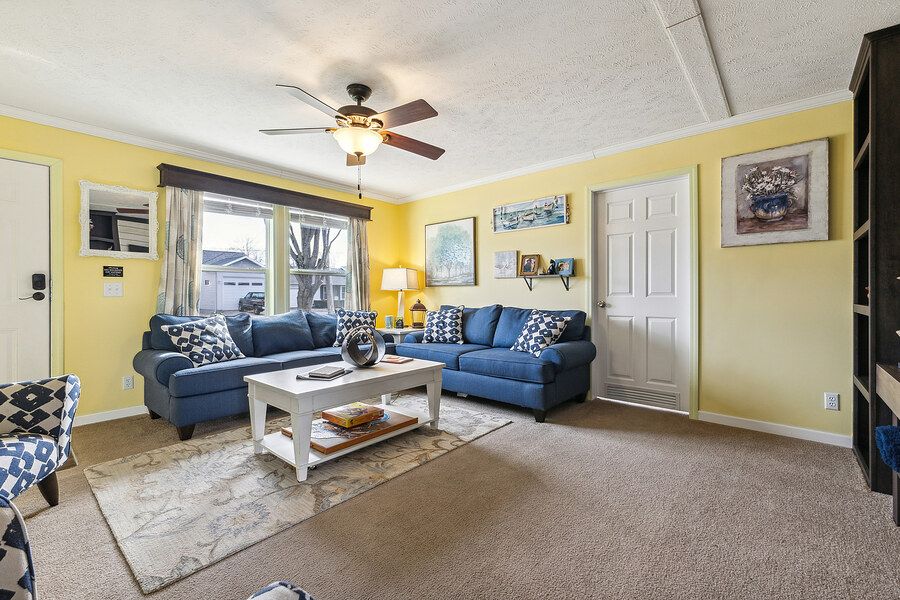 Cozy living room with blue sofas, white coffee table, yellow walls, and carpet flooring under a ceiling fan.