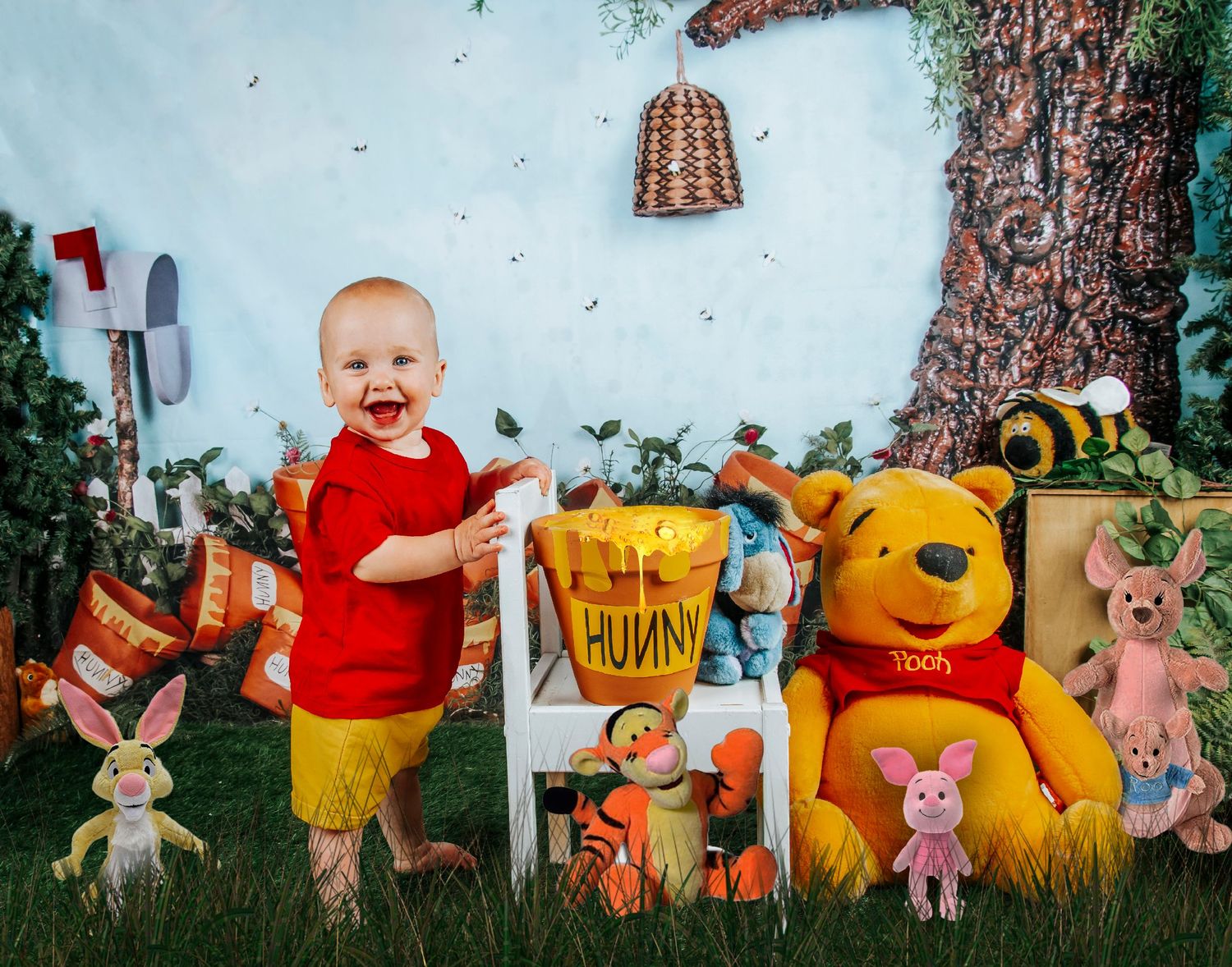 Happy laughing baby in red outfit celebrating first birthday with Winnie the Pooh themed backdrop.