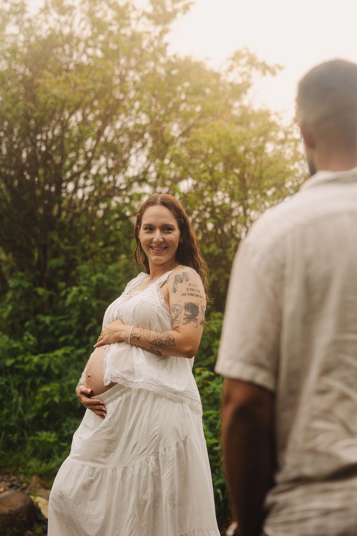 Smiling pregnant woman in white dress with tattoos cradles her baby bump outdoors with lush green trees behind her.