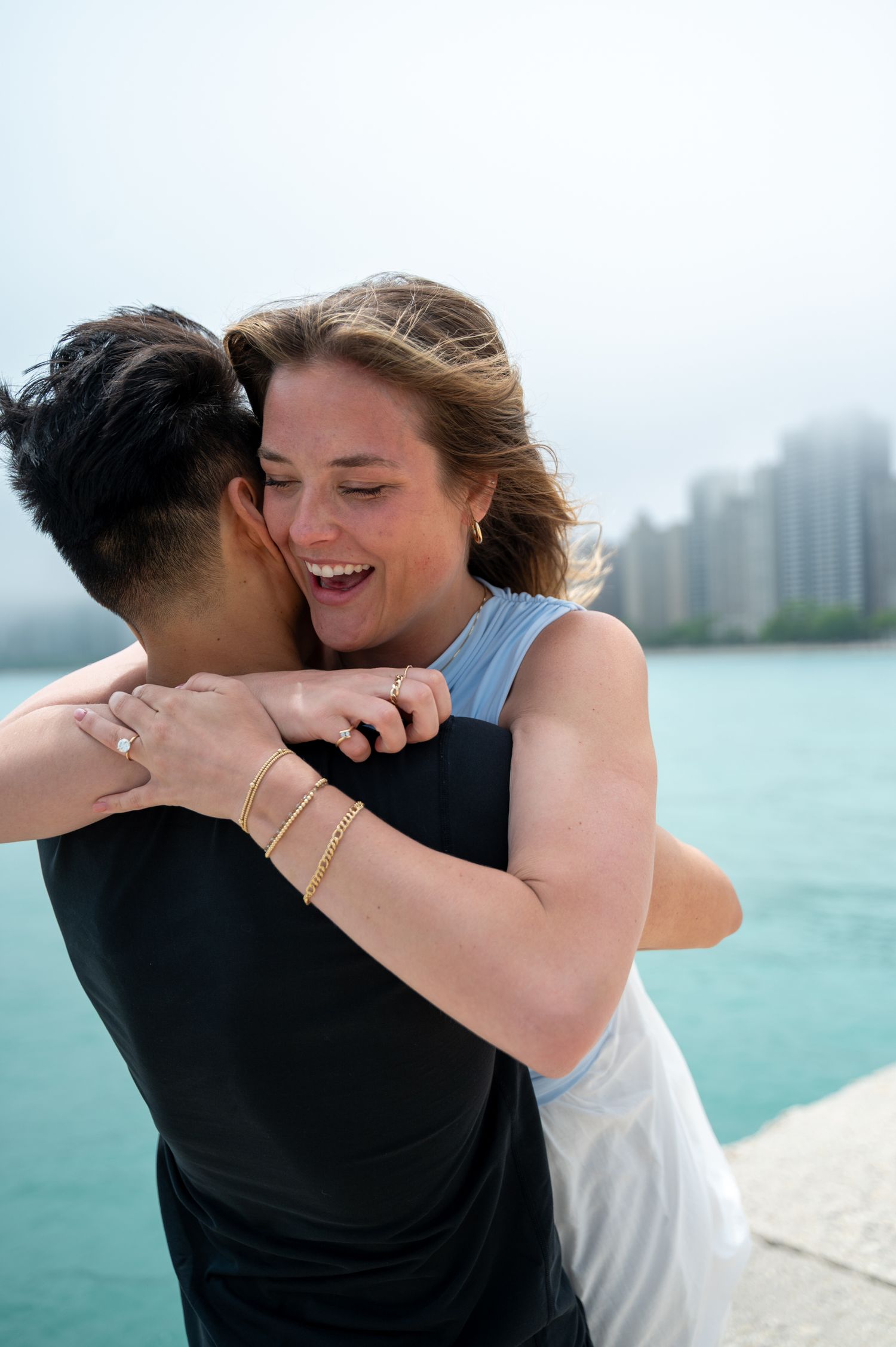 Happy couple embracing outdoors near waterfront, woman laughing joyfully wearing gold bracelets and ring.