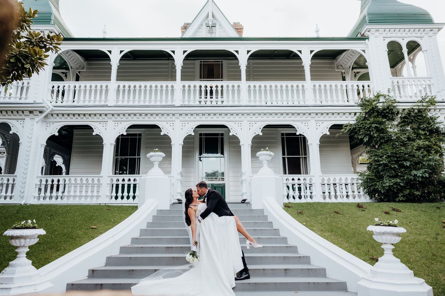 Bride and groom sharing a romantic dip kiss on white stairs of elegant Victorian mansion wedding venue.