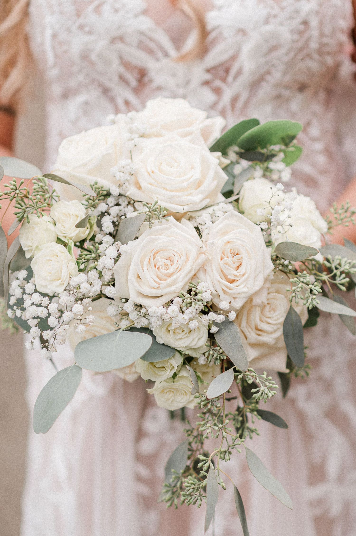 Elegant bridal bouquet featuring white roses, baby's breath, and eucalyptus held by a bride in a lace gown.
