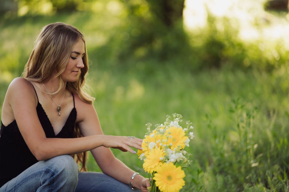 Blonde woman in black top and jeans arranges yellow and white wildflower bouquet outdoors.