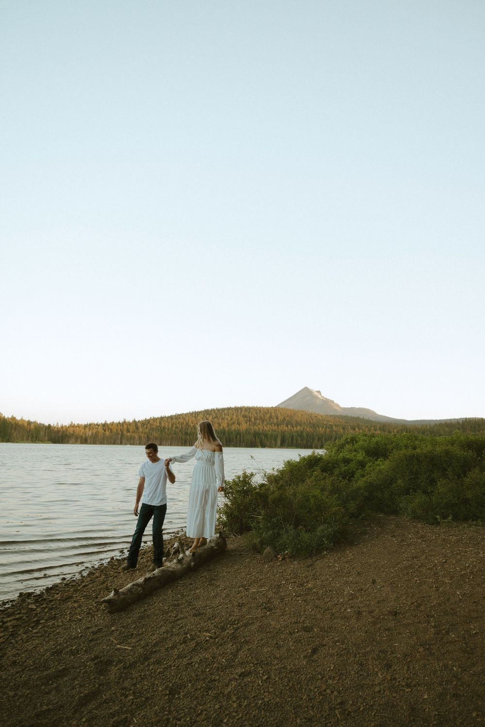 Couple holding hands by a mountain lake shore, woman in white dress, scenic forest and peak backdrop.