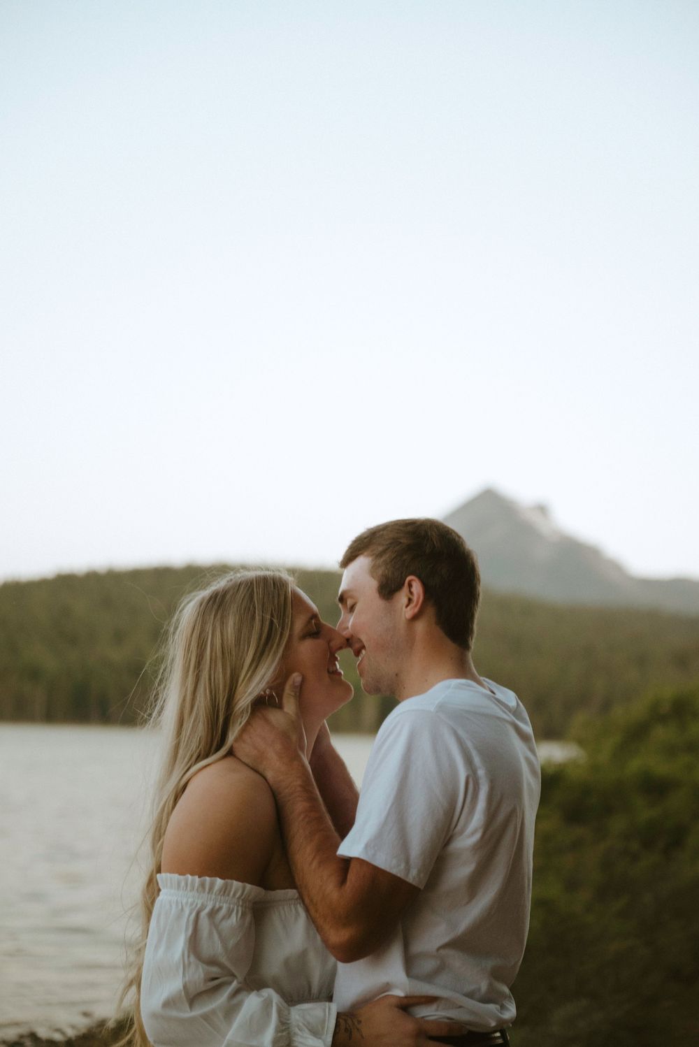 Couple sharing a romantic almost-kiss outdoors near a lake with a mountain backdrop in soft natural light.