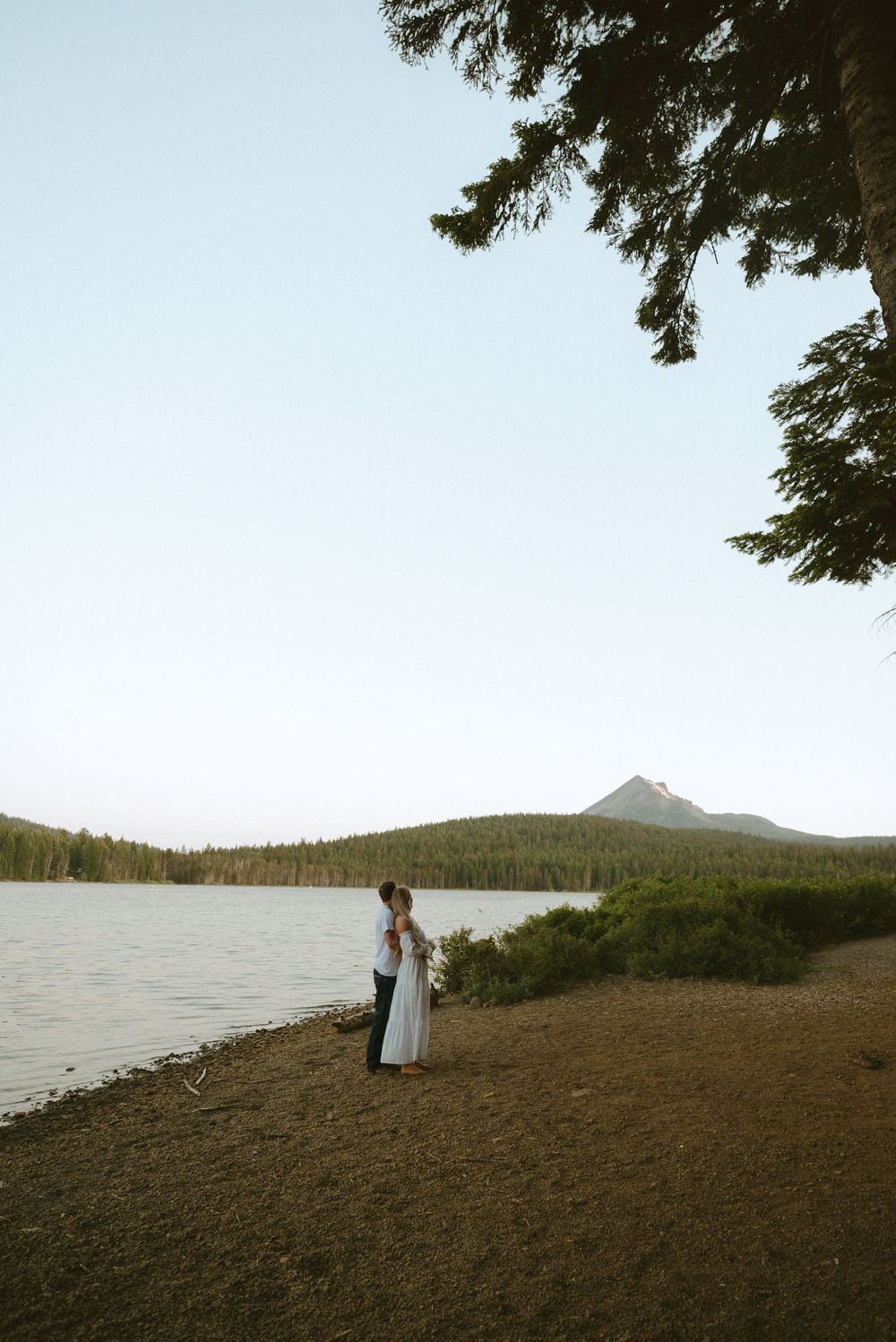 Bride and groom embrace by a serene mountain lake surrounded by pine trees at dusk.