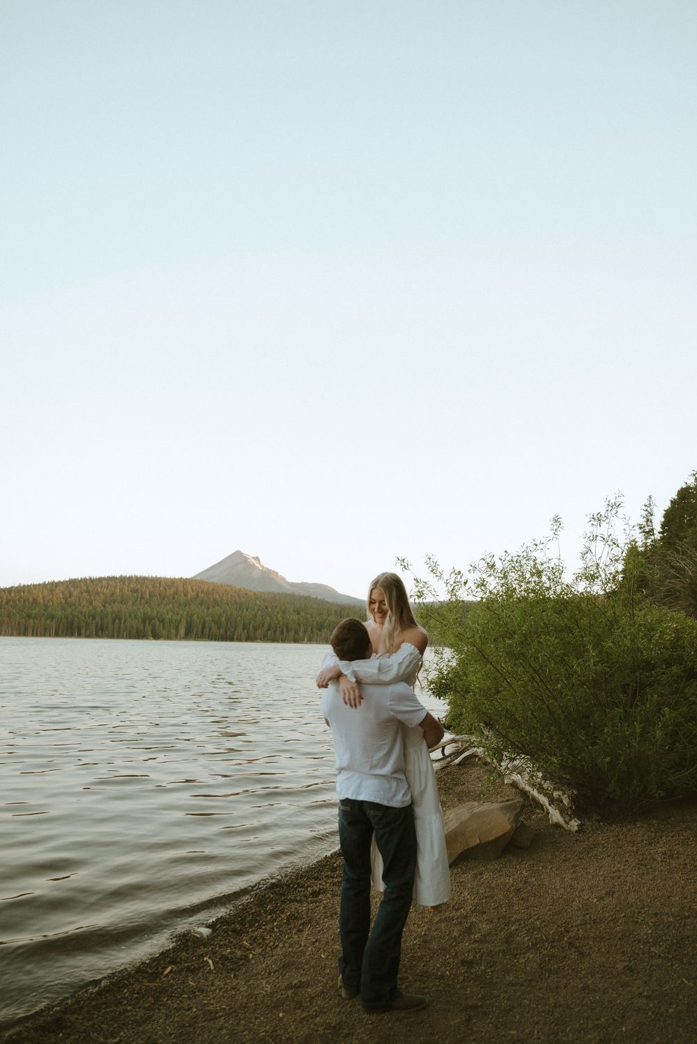 Couple embracing by a serene mountain lake at dusk, man lifting woman in white dress on a sandy shore.