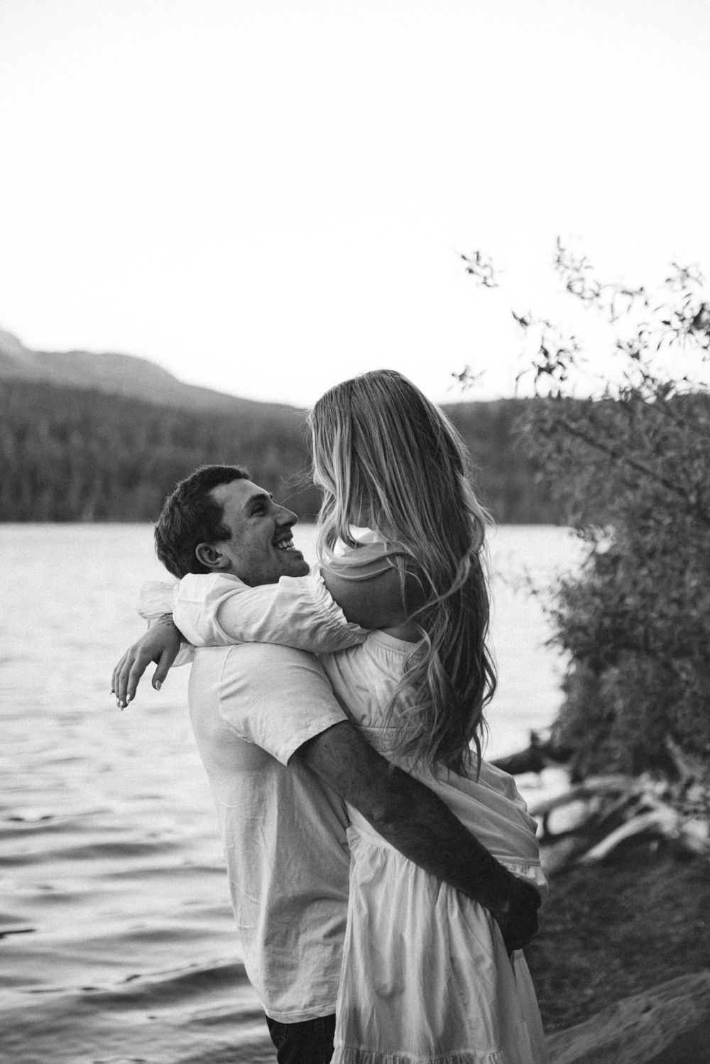 Black and white photo of a couple embracing and laughing by a serene lake with mountains and trees in the background.
