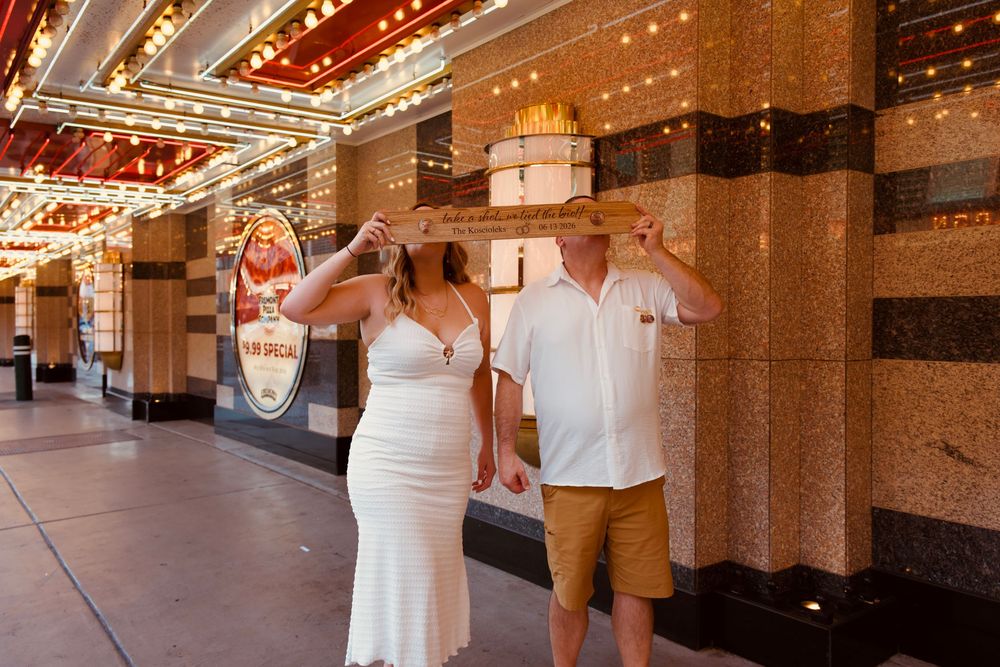 Newlyweds holding a wooden sign together outside a Las Vegas casino under glowing marquee lights.