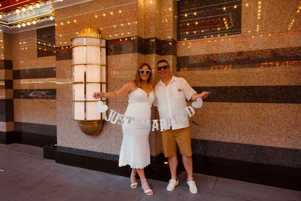 Newlyweds holding a Just Married banner outside a Las Vegas venue with marquee lights.