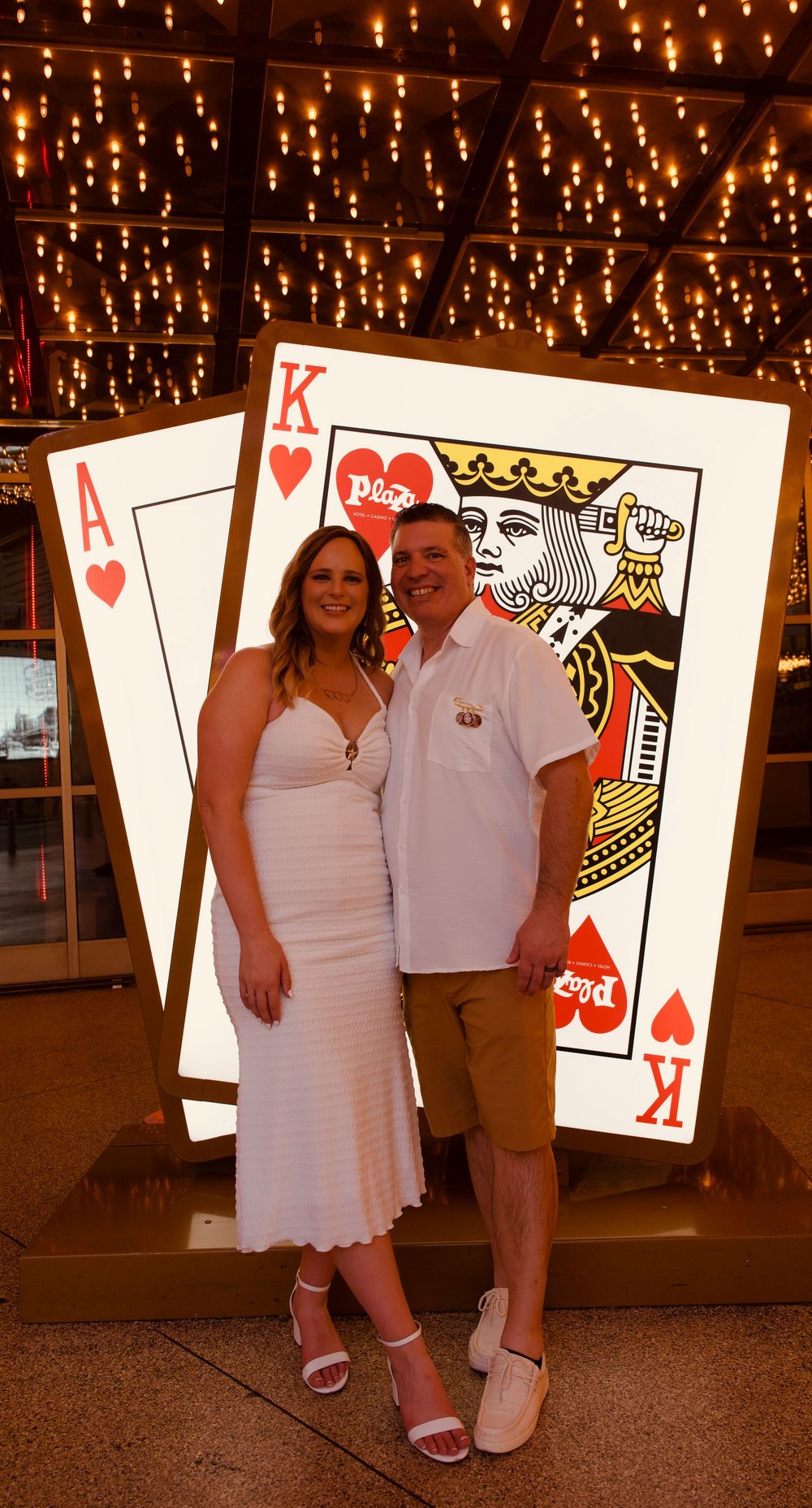 Couple in white outfits posing in front of giant illuminated playing cards at a Las Vegas casino.
