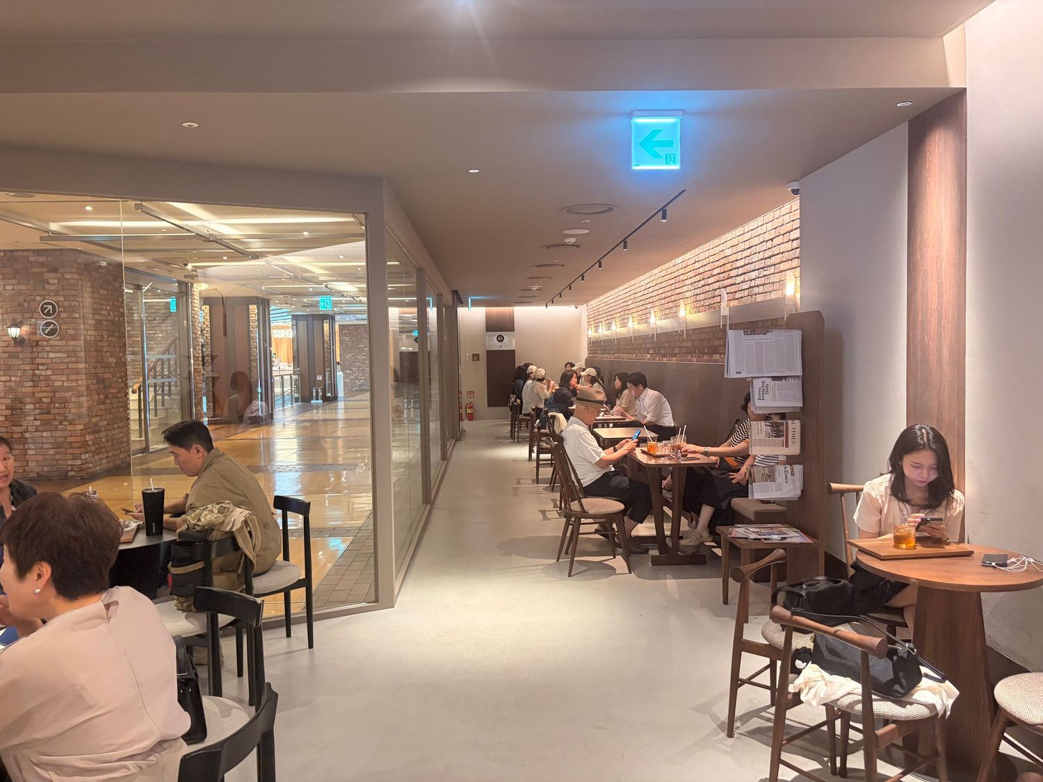 Modern cafe interior with patrons dining at wooden tables along a well-lit corridor with glass walls.