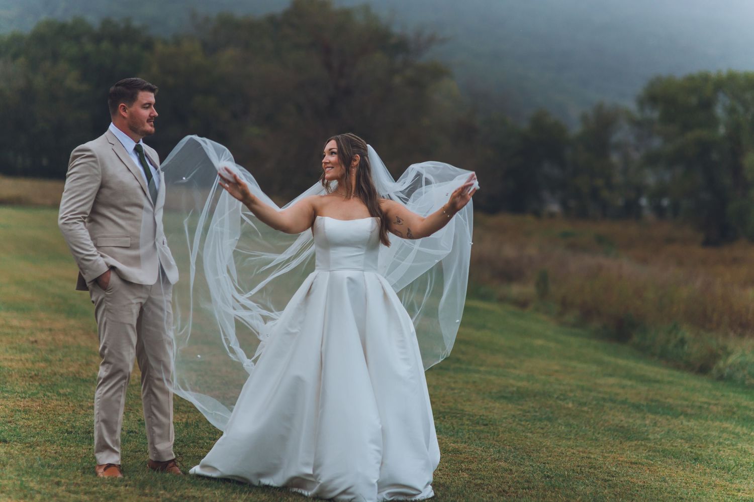 Bride in white ballgown holds flowing veil as groom in tan suit smiles outdoors on lush green field with misty mountains.
