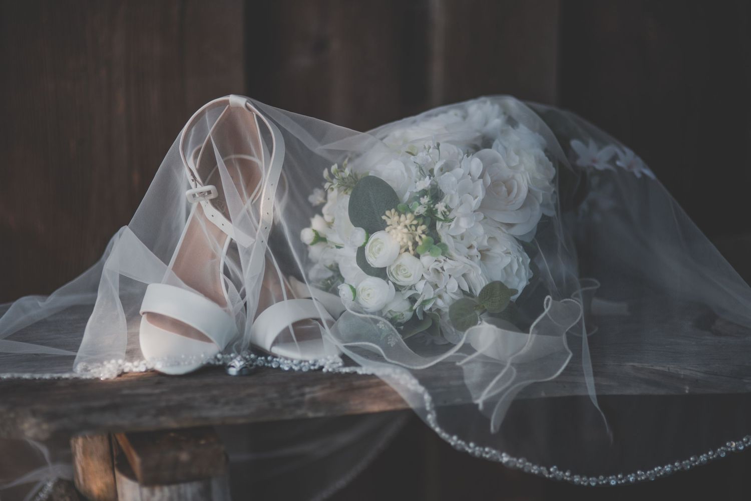 Elegant wedding flat lay featuring white heels, bridal veil, and a white floral bouquet on a rustic wooden surface.