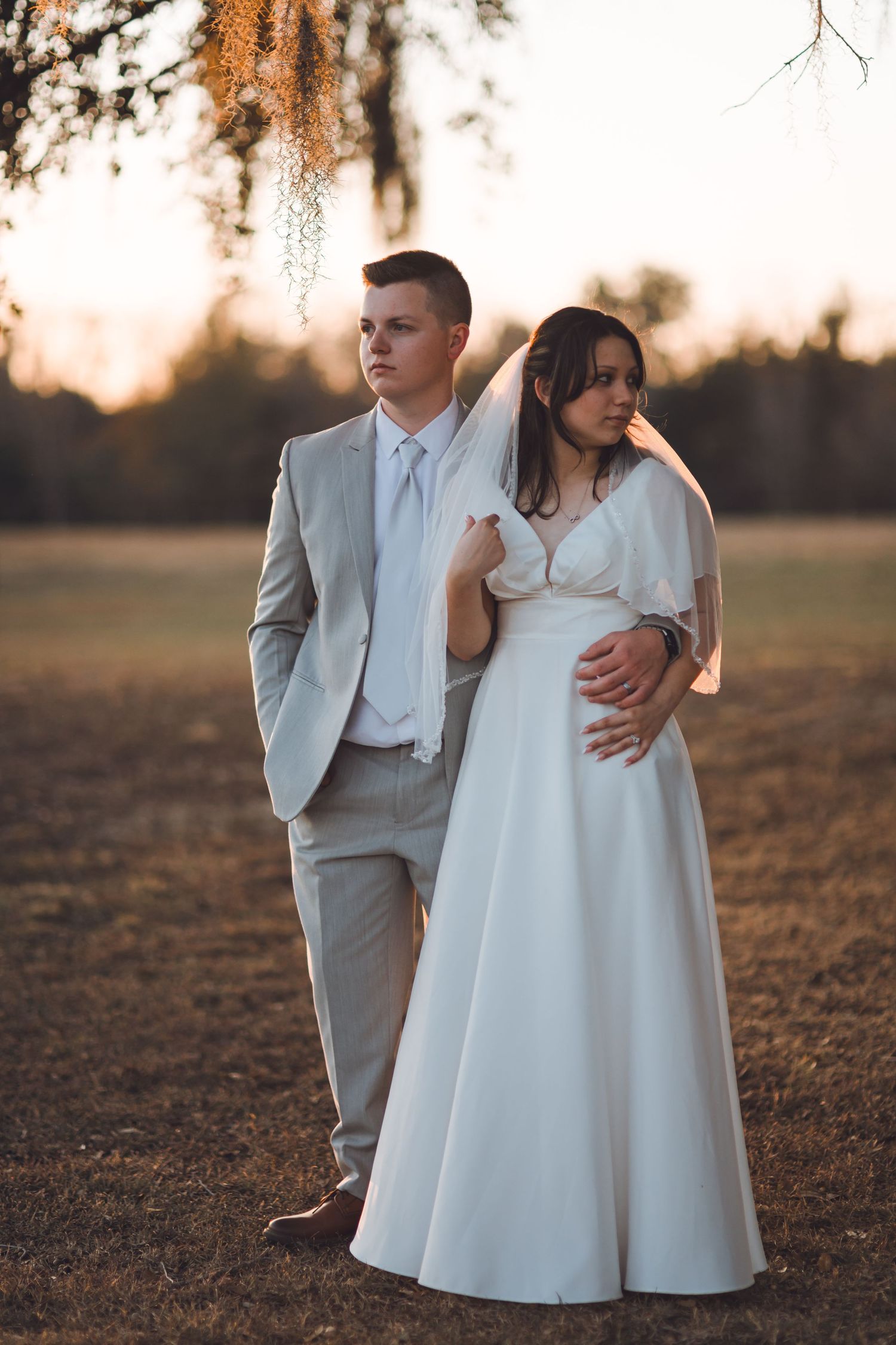 Bride and groom posing outdoors at golden hour, she in a white gown with veil, he in a light gray suit.