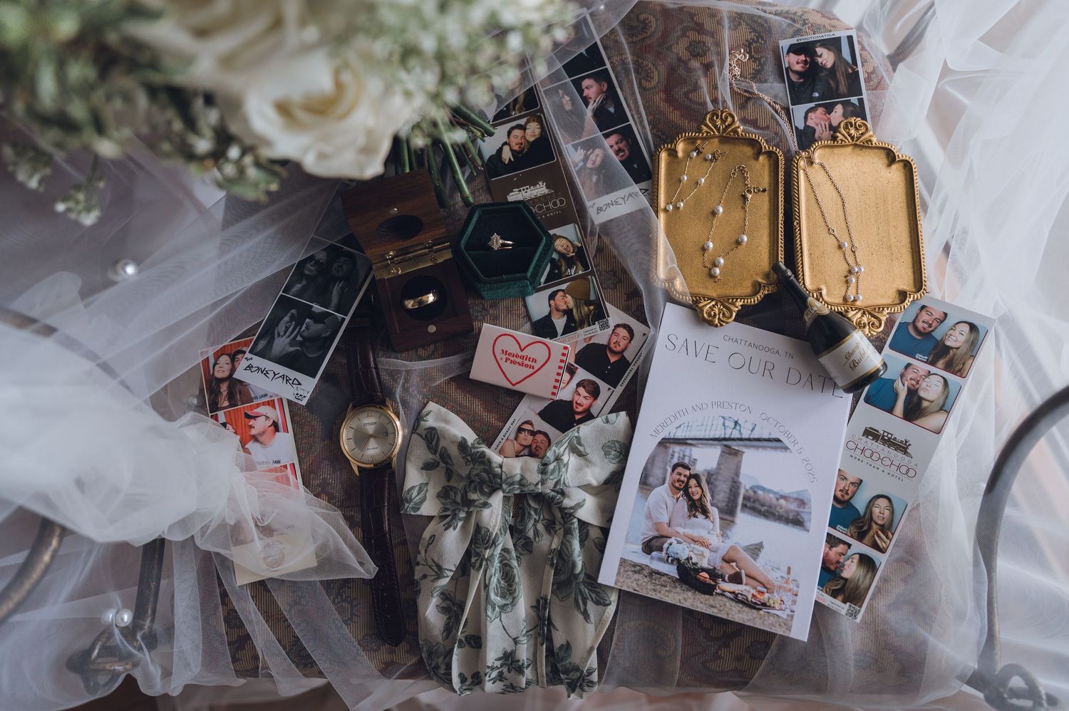 Flat lay of wedding details including photo booth strips, jewelry, perfume bottles, and floral arrangements on a white surface.