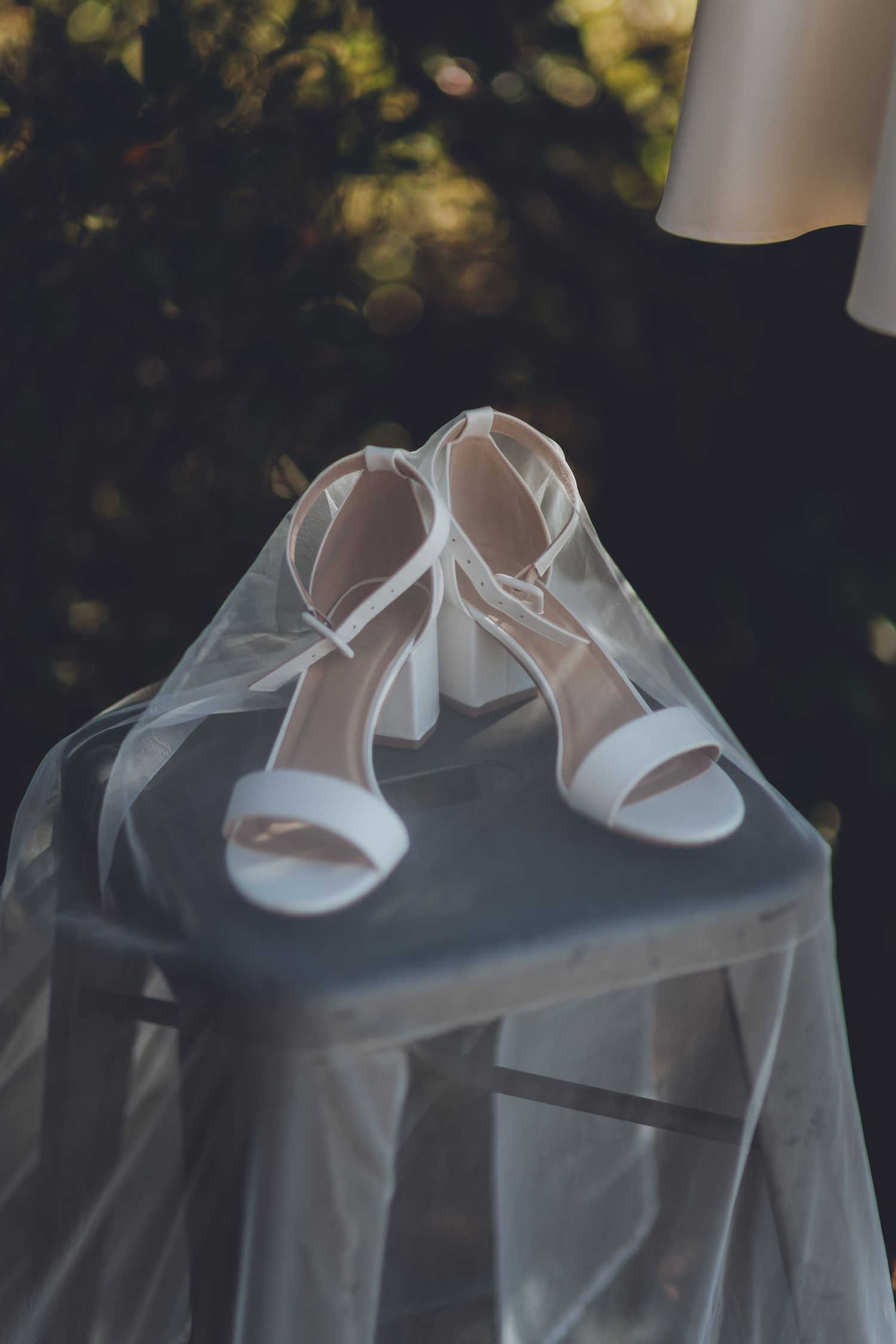 White bridal heels with ankle straps draped in sheer veil on a chair, elegant wedding day accessories.