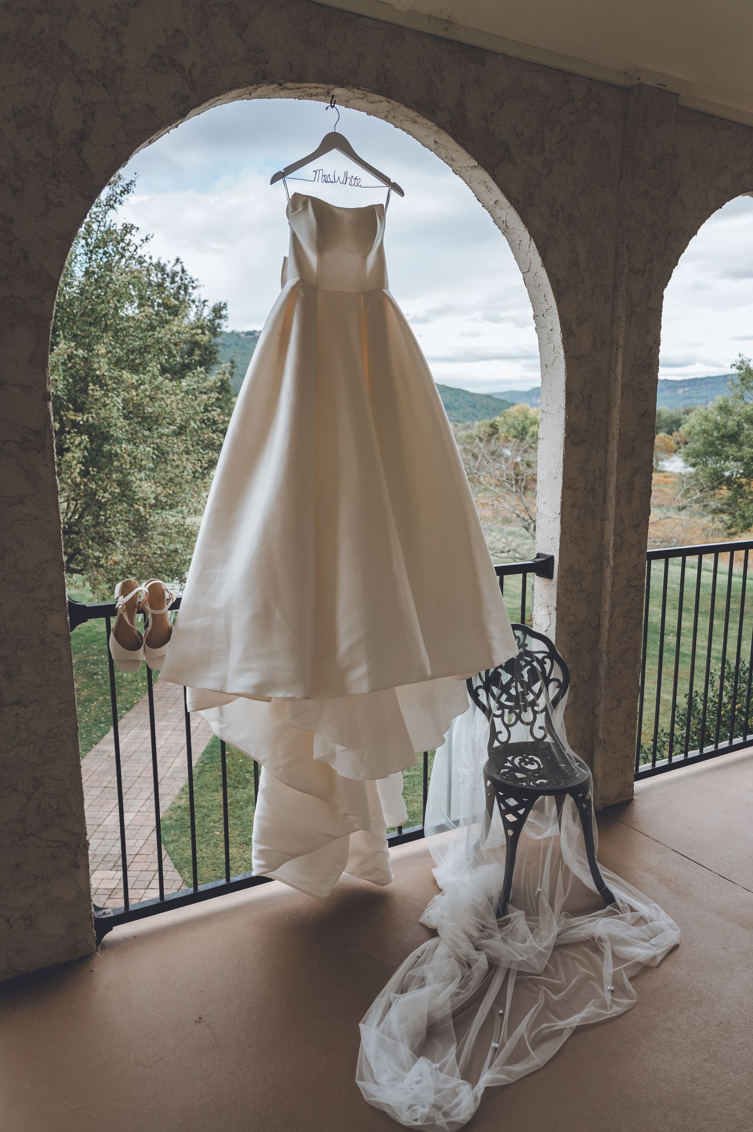 Elegant ivory ball gown wedding dress hanging on personalized hanger on stone arch balcony with scenic mountain views.