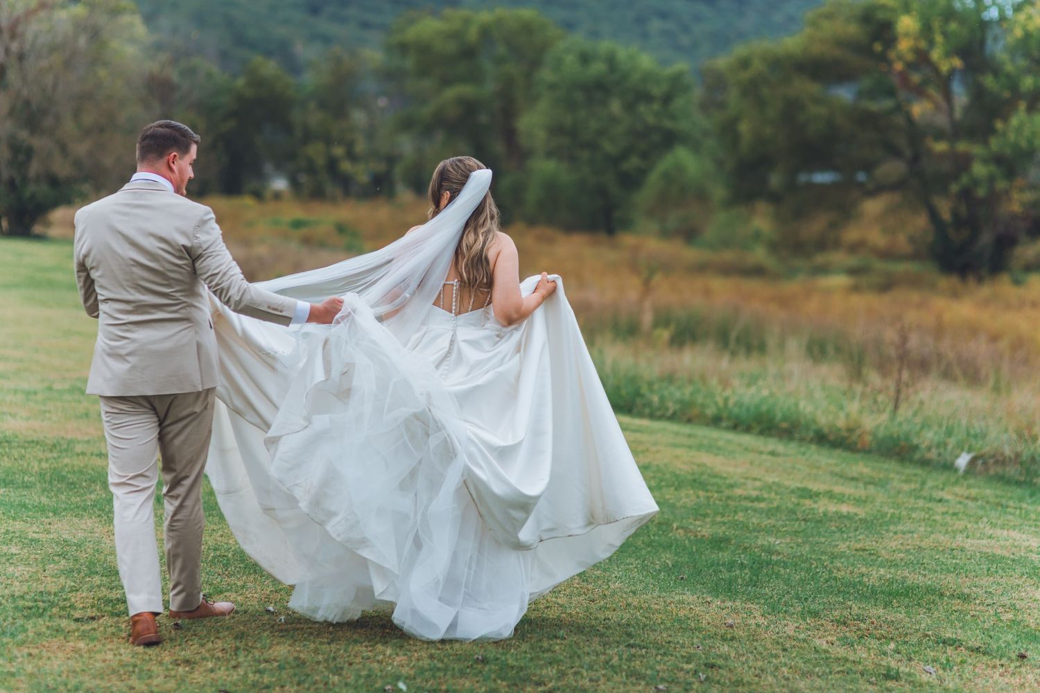 A groom in a beige suit lifts his bride's flowing white wedding veil outdoors on a lush green lawn.