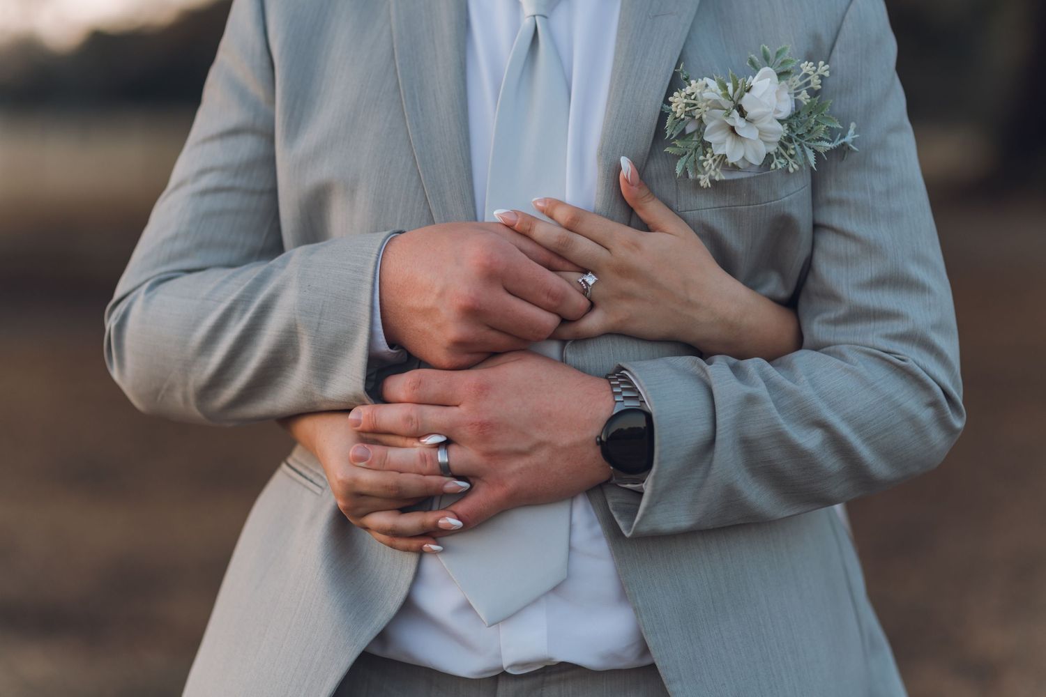 Bride and groom holding hands showing wedding rings, groom in gray suit with white floral boutonniere.