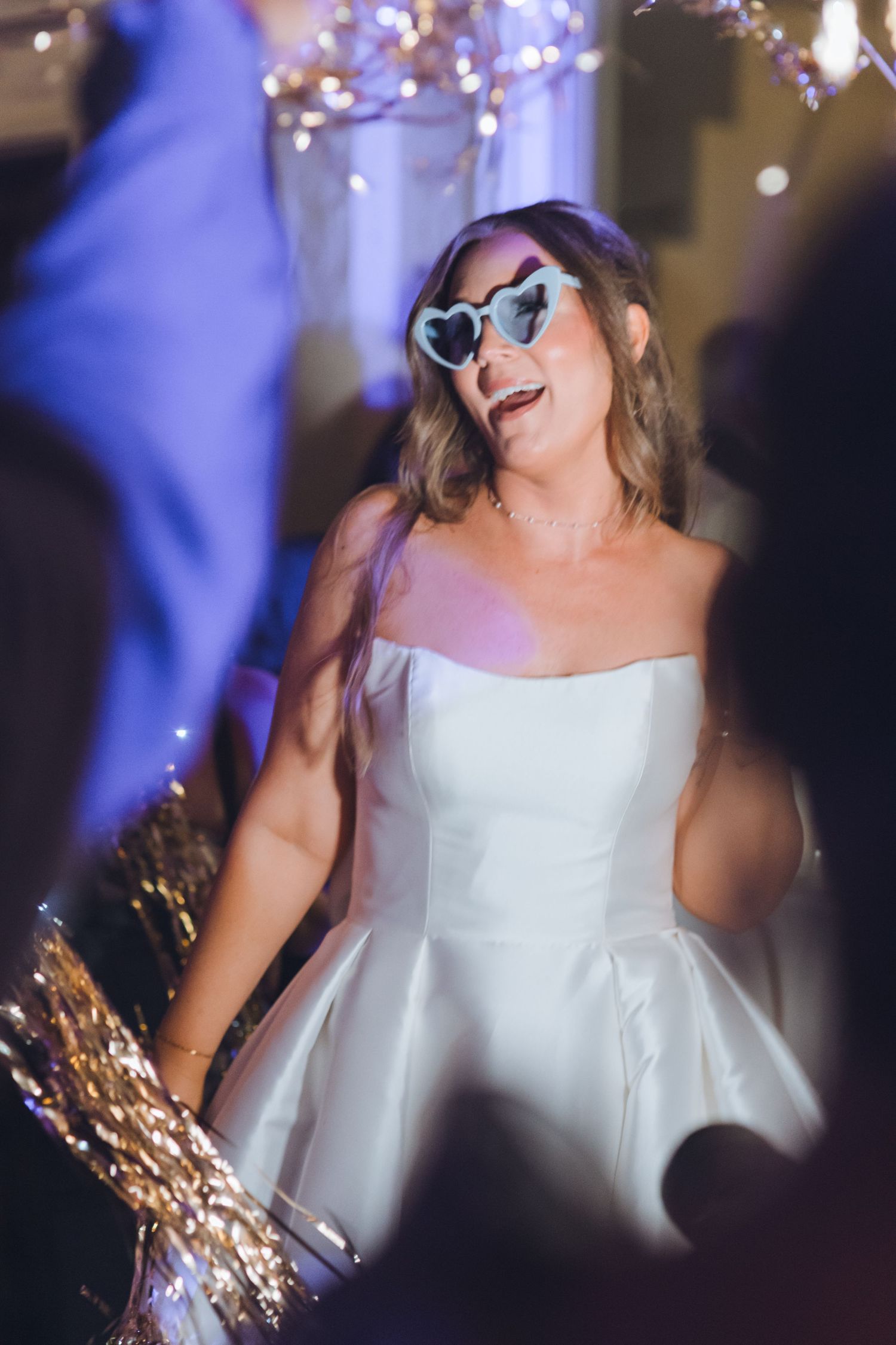 Bride in white strapless gown wearing heart-shaped sunglasses dancing joyfully at wedding reception with bokeh lights.