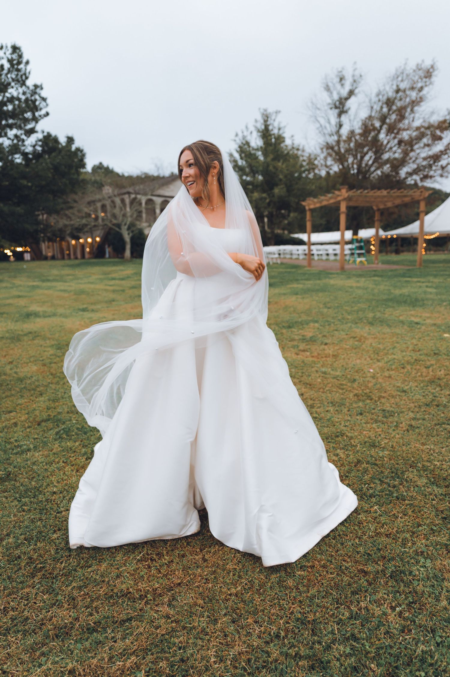 Bride in a flowing white ball gown and veil twirling on green lawn at outdoor wedding venue at dusk.