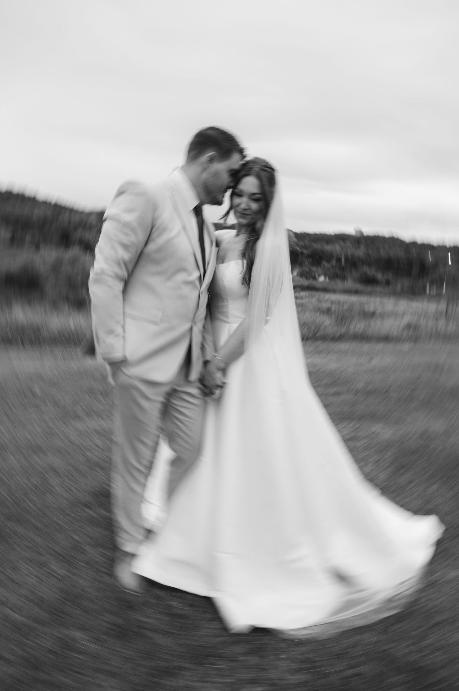 Black and white photo of a bride and groom holding hands, smiling and walking outdoors on their wedding day.